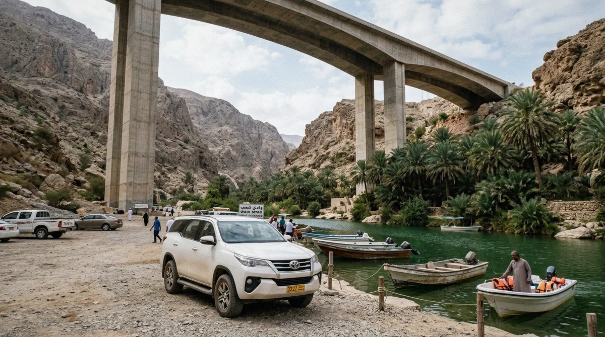 A rental SUV parked beside the Wadi Shab car park under the highway bridge, small boats at the water's edge