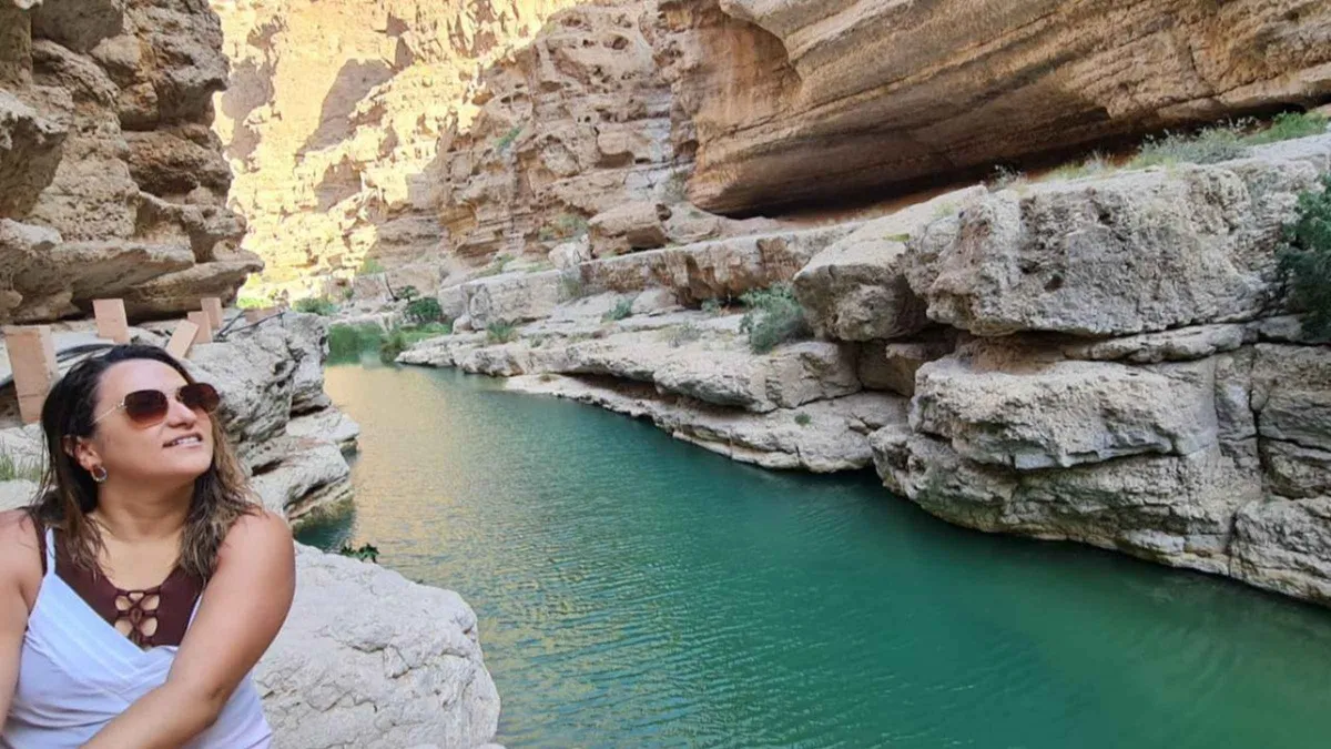 A traveler in sunglasses sitting on a rock above a green pool in Wadi Shab